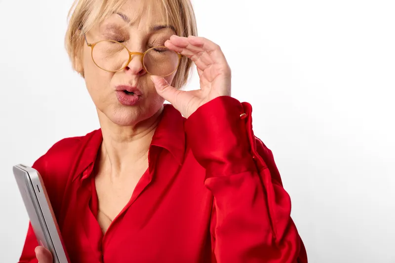 Woman with glasses adjusting near eye, vision and aging, technology using smartphone, eyewear, squinting with discomfort, red blouse on white background, soft studio light care