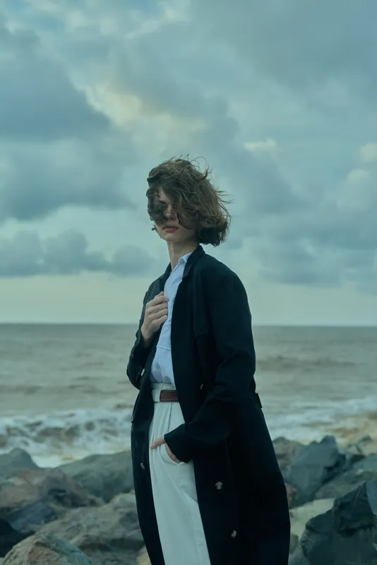 Windy seaside scene with a long dark coat on a rocky shore, windblown hair, moody sky, calm ocean, fashion mood, solitary moment, dramatic lighting, subtle storytelling