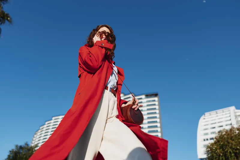 A woman in a bold red coat walks outdoors in an urban setting under a clear blue sky, showcasing street style, confident fashion, and dynamic movement