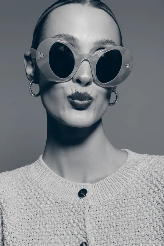 Young white woman with pale skin wearing large round sunglasses and hoop earrings poses with puckered lips in studio portrait. Fashion, style, and beauty concept.