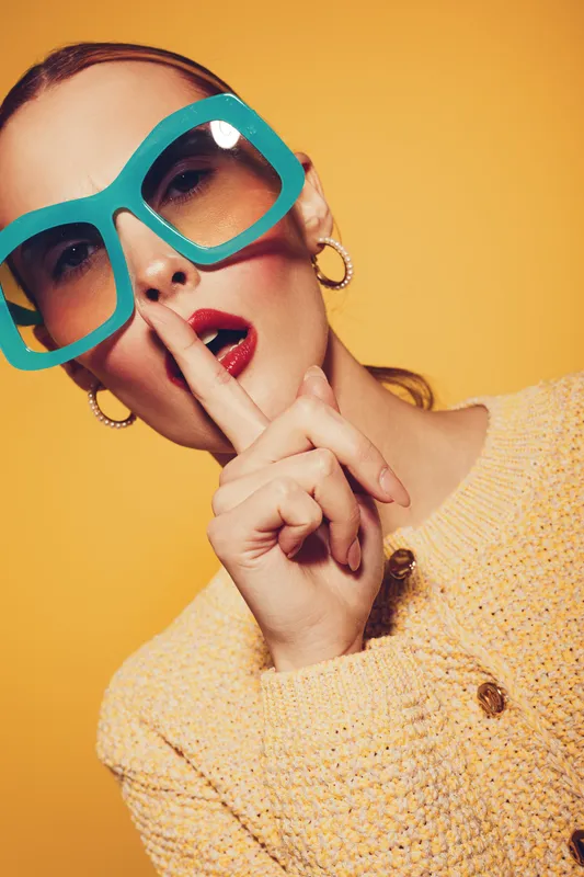 Stylish woman in large turquoise sunglasses and yellow textured jacket makes shushing gesture with finger on lips. Bright yellow background creates bold and fashionable portrait concept.