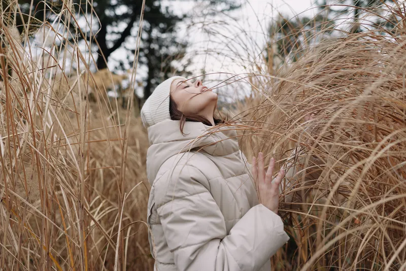 Woman in a warm coat and knit cap among tall grasses, embracing nature and cold air in a serene outdoor portrait, with autumn light.