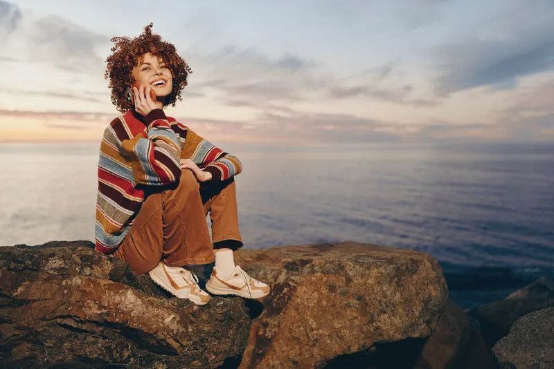 Woman lifestyle at sunset on rocky seaside, smiling while seated on rocks. She wears a scarf and sweater, curly hair, warm evening light, ocean horizon in view, peaceful outdoors travel mood.