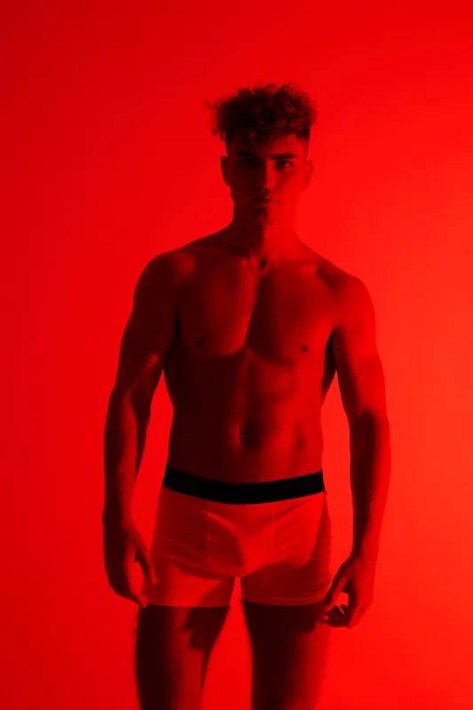 Male model wearing underwear posing on red background shirtless with muscular fit body and confident posture spotlight looks serious athletic fashion studio lighting.