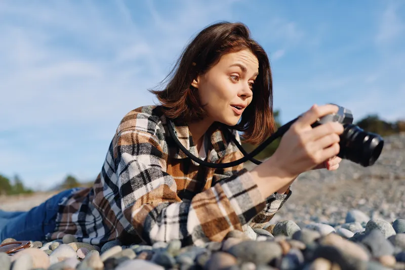 Woman photographer lying on pebbles beside a sunny beach, outdoors and focused through a camera for a candid shot, capturing natural light and coastal details