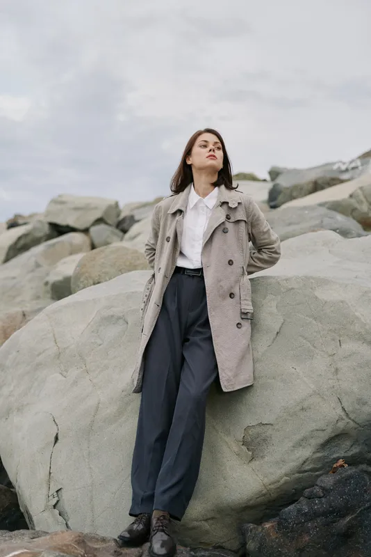 Young woman in gray coat leans on large rocks outdoors under cloudy sky. She wears white shirt and dark trousers, looking confident and relaxed. Concept of fashion and nature.