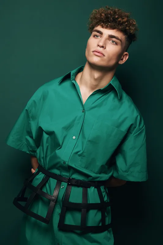 Young man with curly hair wearing green oversized shirt and black harness belt posing against dark green background in studio. Fashion model with serious expression and modern style.
