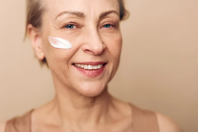 Smiling mature woman with light skin applies white face cream on cheek, posing against beige background. Concept of skincare, antiaging routine, beauty care, and healthy lifestyle for seniors.