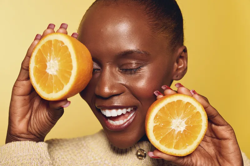 Joyful African woman with dark skin holds two orange halves near her face, smiling with closed eyes. Concept for natural beauty, healthy lifestyle, and vitamin C benefits.