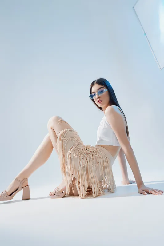 Young woman in stylish sunglasses and a fringed skirt poses seated on a white floor in a bright studio. Concept of woman beauty fashion with modern elegance and confidence.