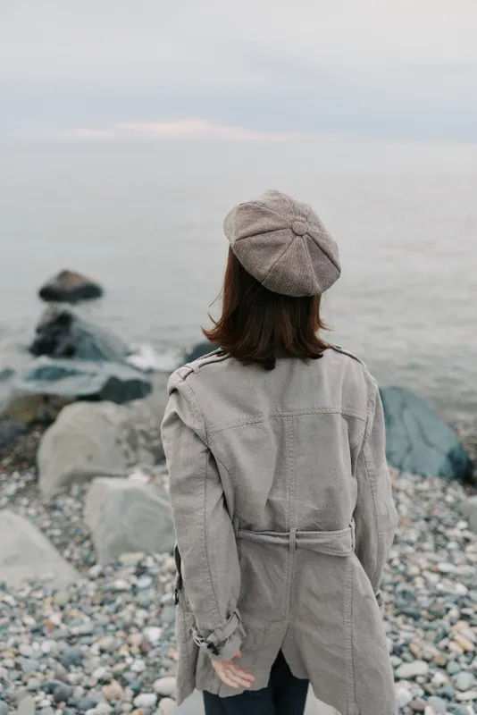 Woman in checkered coat and beret stands on rocky shore looking at calm sea under cloudy sky. Concept of autumn, solitude, and peaceful nature moment.