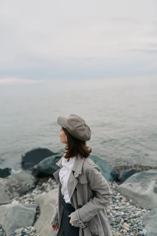 Young woman in vintage coat and hat stands on rocky shore looking at calm sea. Overcast sky creates peaceful mood. Concept of solitude, nature, and thoughtful moment outdoors.