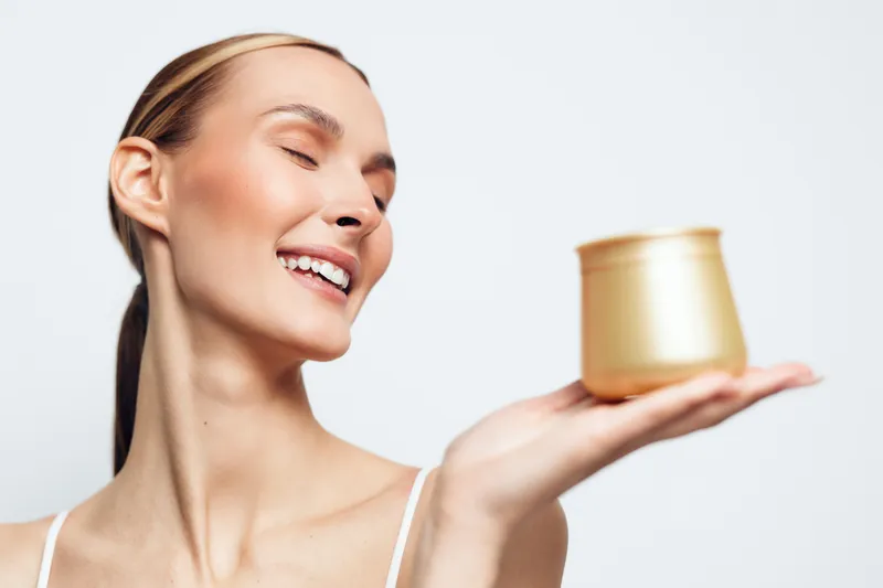 A happy young woman with clear skin smiles with closed eyes while holding a golden cosmetic jar on her palm against a white background. Concept for beauty skincare and wellness.