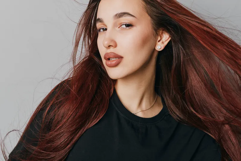 Woman with long red hair wearing black shirt posing with flowing hair on gray background. Portrait of confident female with natural makeup and earrings in studio lighting.