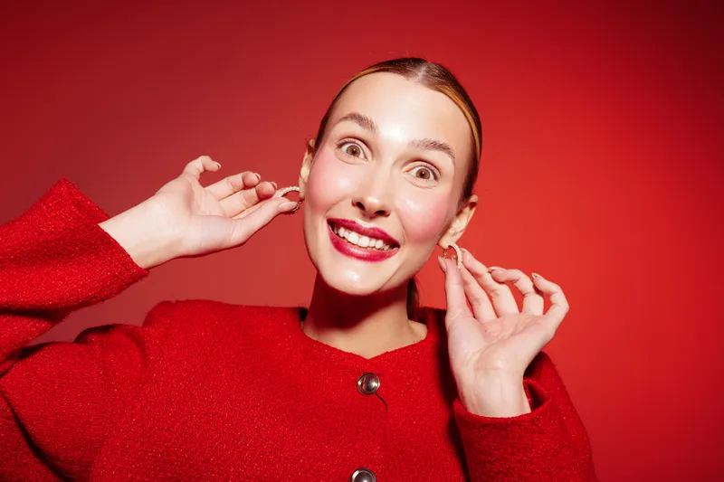 Smiling young woman in red sweater adjusts earrings on red background. Bright makeup and joyful expression create festive mood. Concept for fashion, beauty, and happiness.