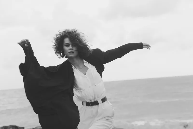 Woman wind coat sea movement monochrome fashion outdoors ocean expression, a dynamic figure dances along the shore with flowing fabric and a dramatic horizon
