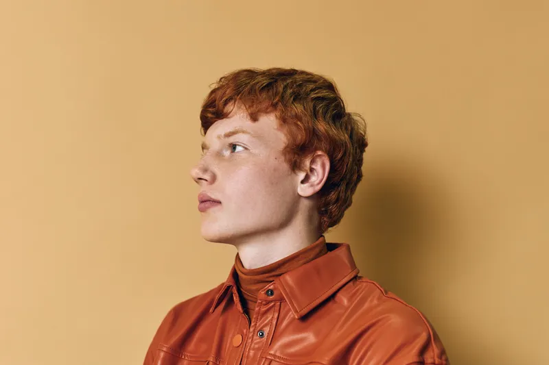 Young man with red hair wearing brown leather jacket posing in profile on beige background indoors for fashion, style, or portrait photography with natural lighting.