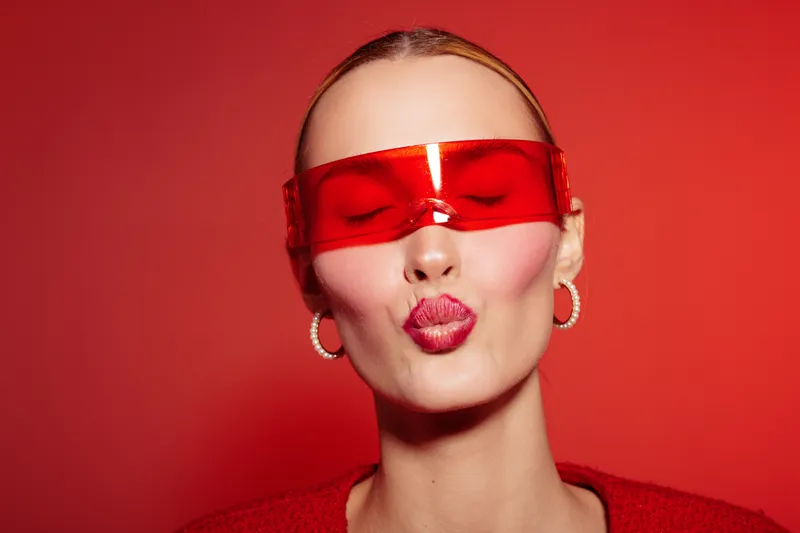 Young woman with red lips and red visor makes a kiss face on red background. Fashion portrait with hoop earrings and smooth skin. Concept of beauty, style, and modern accessories.