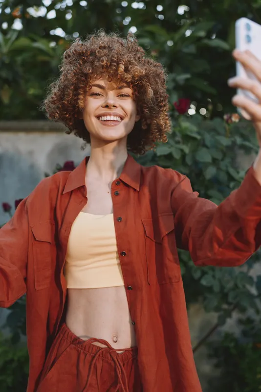 A cheerful woman takes a selfie outdoors, wearing a rust jacket and crop top, capturing casual lifestyle and fashion vibes in natural light