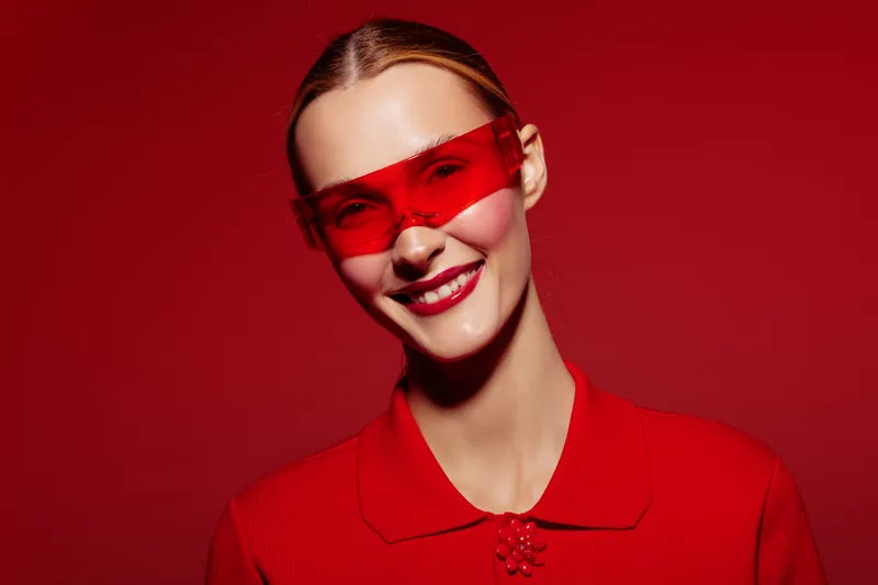 Smiling woman in red sunglasses and red outfit poses against red background. Fashion portrait with vibrant colors and confident expression. Concept of style and modern look.