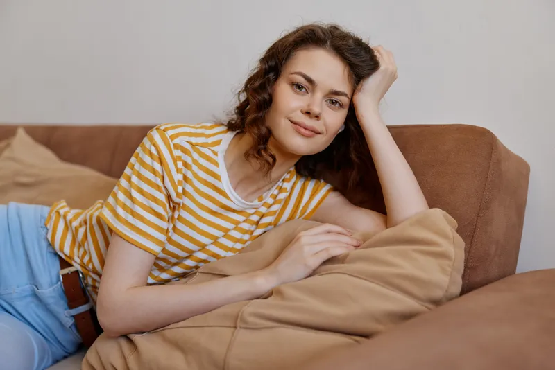 A relaxed woman lounges on a sofa at home, wearing a striped tee and blue jeans, smiling softly. Cozy living room scene conveying casual comfort and everyday downtime.
