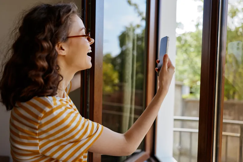 Happy woman in a striped shirt enjoys a bright home moment, taking a selfie by the window with a phone, showcasing cheerful, casual lifestyle indoors. Natural light, relaxed atmosphere.