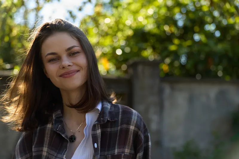 Young woman outdoors in a plaid shirt smiles softly, enjoying warm natural light in a sunlit garden, conveying a relaxed lifestyle mood