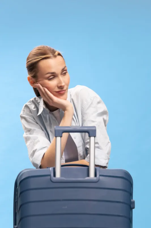 A young woman leans on a blue suitcase with a tired expression, wearing a white shirt against a blue background. Concept of travel fatigue, waiting, and journey anticipation.