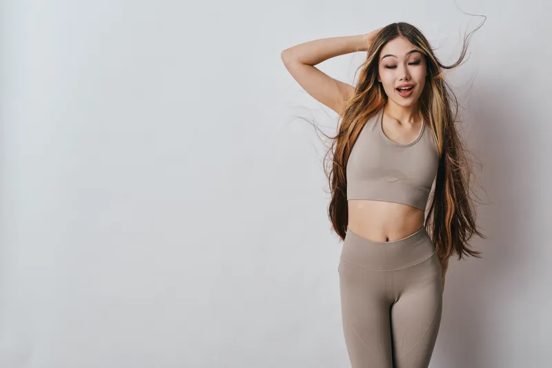 Young woman in sportswear with long hair posing energetically against a light wall. Fitness, health and lifestyle concept with casual outfit and joyful expression indoors.