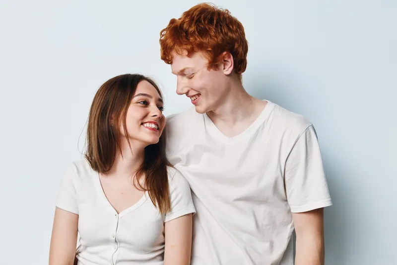 Young couple, smiling and looking at each other, wearing casual white t shirts against plain light background. Friendship, happiness and connection between man and woman.