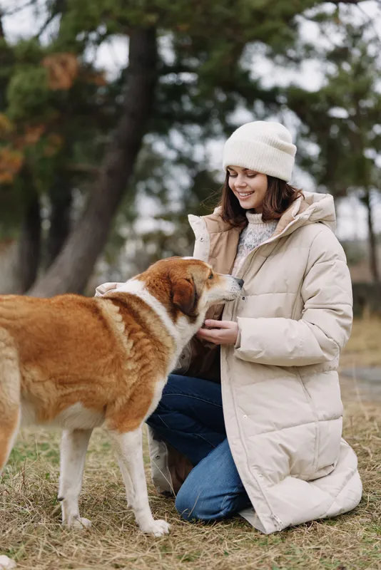 Dog and woman share a gentle moment outdoors in a light winter coat and knit hat, kneeling on grass as they interact with friendly eye contact and trust, and warmth.