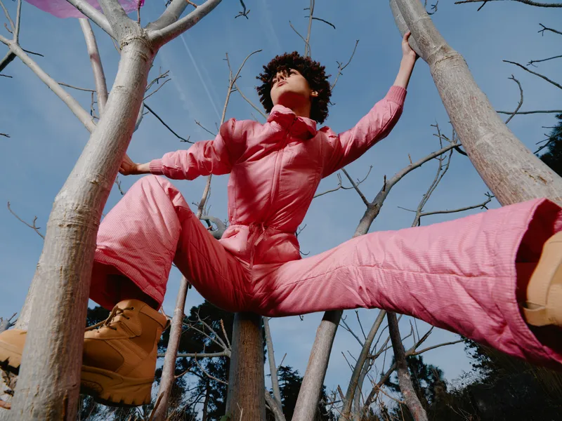 Outdoor athlete climbing trees in a pink jumpsuit creating a dynamic pose among branches under blue sky, capturing balance, freedom and energy in motion and strength