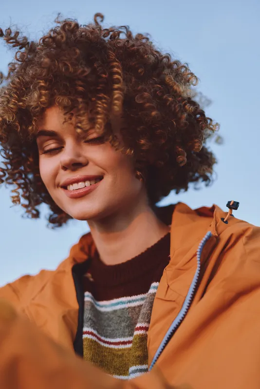 Smiling woman with curly hair outdoors, wearing a tan jacket and striped sweater, close portrait against a clear blue sky, warm lifestyle mood, natural sunlight in summer breeze