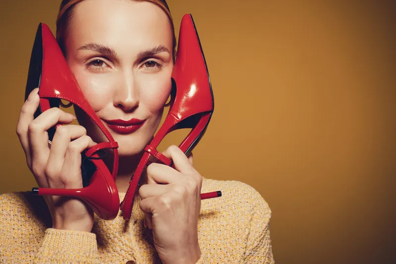 Young woman holds red high heels near face, wearing yellow sweater. Studio portrait with warm lighting on brown background. Concept of fashion, style, and elegance.