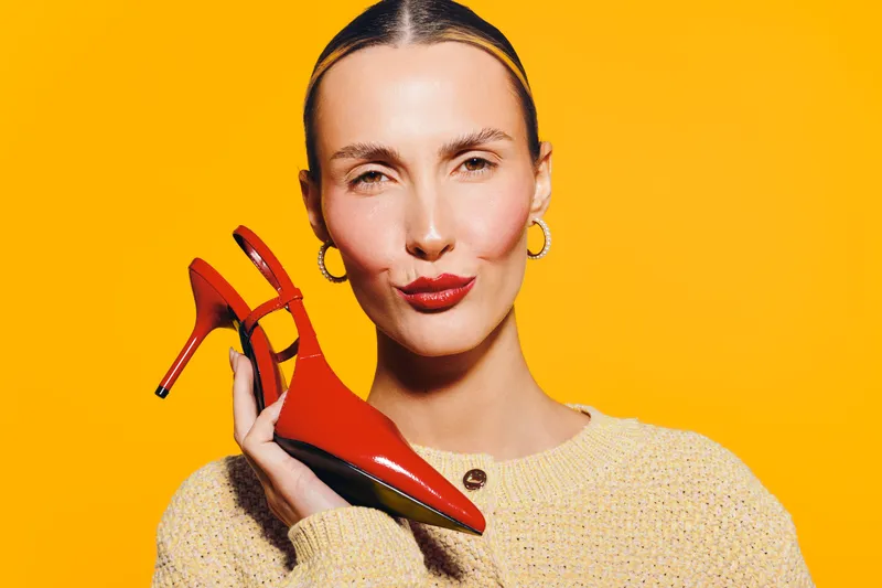 Stylish woman with slicked hair holds red high heel shoe near face on bright yellow background. Concept of fashion, elegance, and modern style with vibrant colors and confident expression.