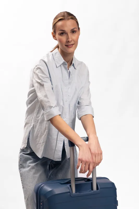 Confident young woman in casual shirt and pants leans on blue suitcase handles, smiling slightly against white background. Concept of travel preparation, tourism, and modern lifestyle.