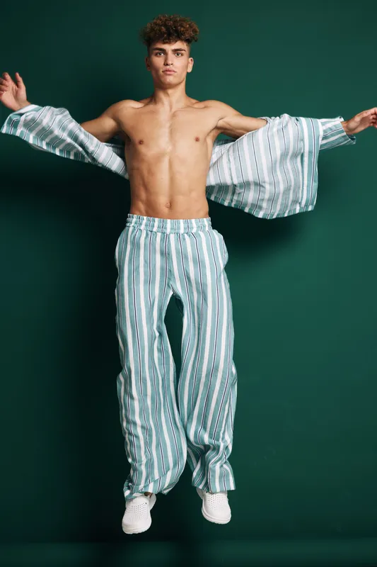 Young man jumping with open striped shirt and matching pants against dark background. Casual fashion, athletic body, confident pose and dynamic movement in studio lighting.