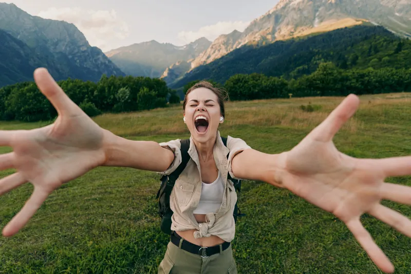 Happy woman with backpack stretching arms towards camera in green meadow with mountains in background. Outdoor lifestyle, adventure, travel, and freedom concept.