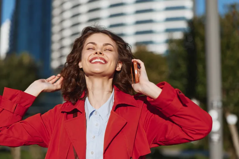 Smiling woman in a red coat outdoors, phone to her ear, sunlit urban background, casual street fashion, joyful mood, candid lifestyle and carefree attitude vibrant energy.