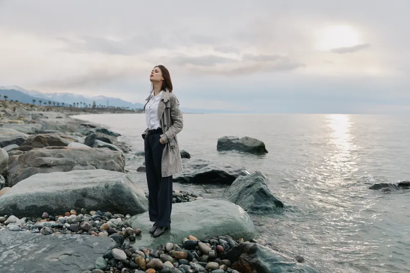 Woman in trench coat stands on rocky shore near calm sea under cloudy sky. She looks peaceful and contemplative, enjoying nature and fresh air by the water. Concept of relaxation and freedom.