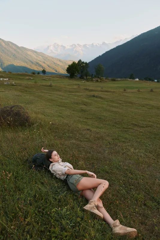 Young woman resting on a sunlit valley meadow with distant mountains, conveying a calm, free mood and a connection to nature.