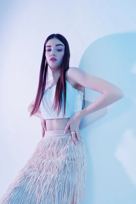 Young woman beauty fashion model poses confidently in stylish white crop top and textured skirt. Studio shot with soft lighting and pastel background. Concept of modern fashion.