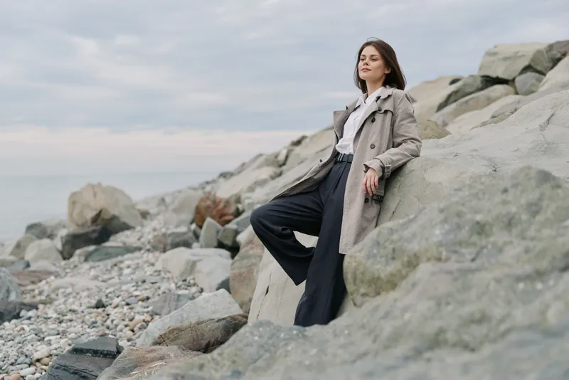 A confident woman in a trench coat stands on rocky shore with cloudy sky background. Concept for fashion, autumn style, and outdoor lifestyle with natural landscape.