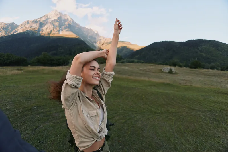 Young woman enjoying outdoor lifestyle, stretching arms in a sunlit grassy field with distant mountains in the background. Cheerful, calm moment conveying wellness, vitality, and freedom