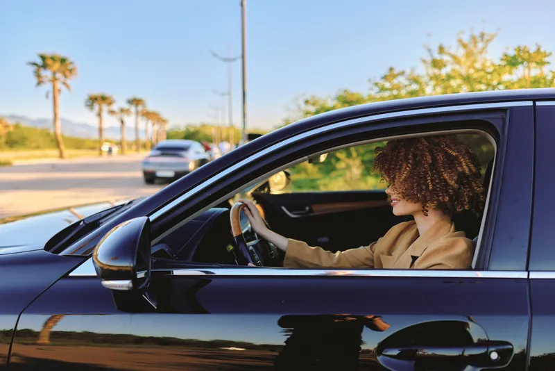 Cheerful woman driver in a car, retro film color tones, sunlight on face, relaxed posture, window down, street scene with palm trees, candid lifestyle moment outdoors