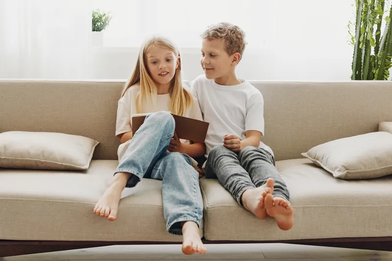 Two children sitting barefoot on a beige sofa, reading a book together in a bright living room with pillows and plants. Concept of childhood, learning, and family leisure time.