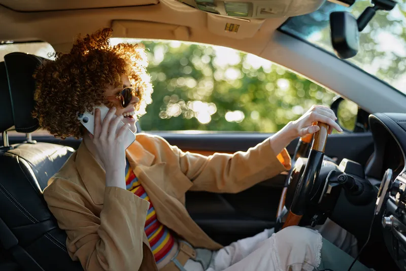 Woman with curly hair driving a car while talking on a mobile phone. Soft warm lighting and retro color tones create a casual lifestyle mood with everyday urban charm.