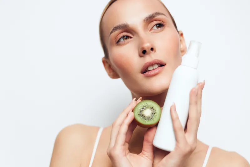 A young woman holds a kiwi beside a white lotion bottle, illustrating a skincare routine. Bright studio lighting highlights healthy skin, hydration, and natural beauty through simple self care