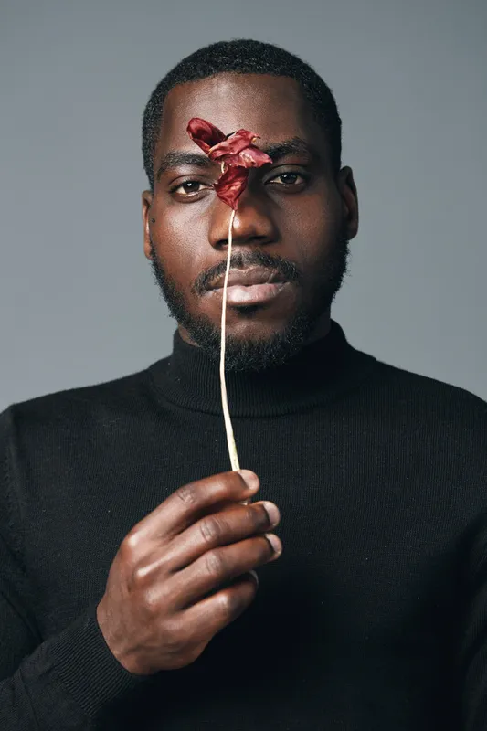 African man with dark skin holding dried flower in front of face in creative conceptual visual style. Portrait with black turtleneck and minimalistic gray background for artistic expression.
