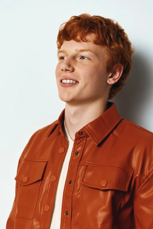 Young man with red hair wearing brown leather jacket smiling and looking aside with white background for portrait and casual fashion style.